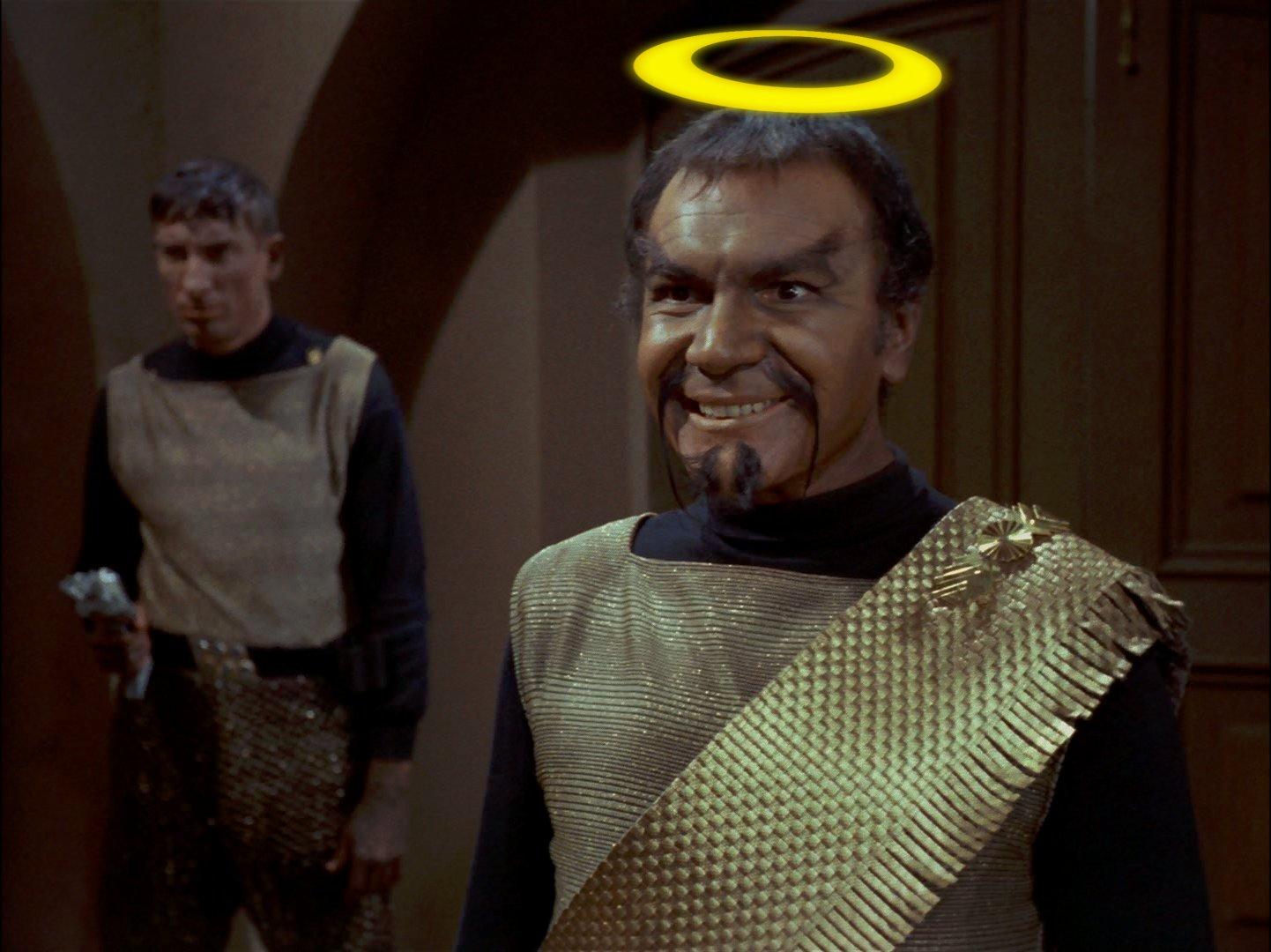 Ein Screenshot aus Star Trek, der den Klingonen Kor (gespielt von John Colicos) zeigt. Er hat braune Haut, kurze, dunkle Haare, zwei lang herunterhängende Schnurrbartspitzen und am Kinn einen gespaltenen Spitzbart, und trägt die TOS-typischen Klingonenuniform (goldenes, ärmelloses Shirt über einem schwarzen, langärmeligen Untershirt) mit goldener Scherbe. Dementsprechend hat er auch keine klingonischen "Stirnwülste". Kor grinst schemlisch-triumphierend. Über seinem Kopf ist ein comic-hafter, goldgelber Heiligenschein. Im Hintergrund steht ein weiterer Klingone in einer ähnlichen Uniform, allerdings ohne Scherpe. Dieser zweite Klingone ist unwichtig.